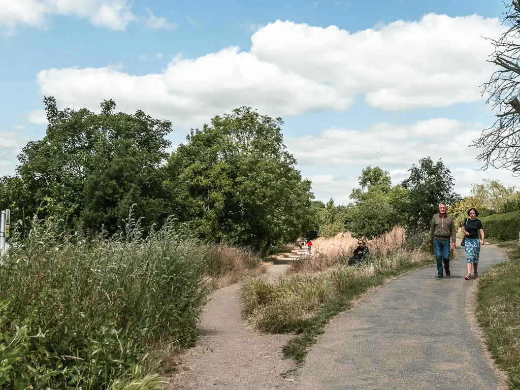 A split in the walking path, with grass between the split. There are bushes and trees lining the left path, and two people walking along the right path.