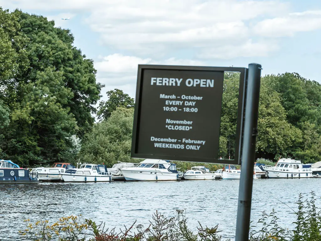 A black sign saying 'Ferry open', with a backdrop of the river, then white boats and trees lining the other side.