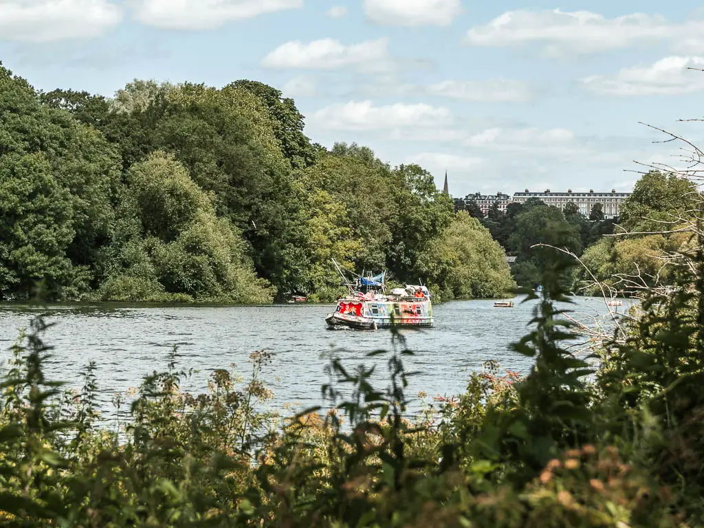 A colourful barge in the middle of the river, on the walk from Hampton Court to Putney. The river is lined with very large green leafy trees on the other side.