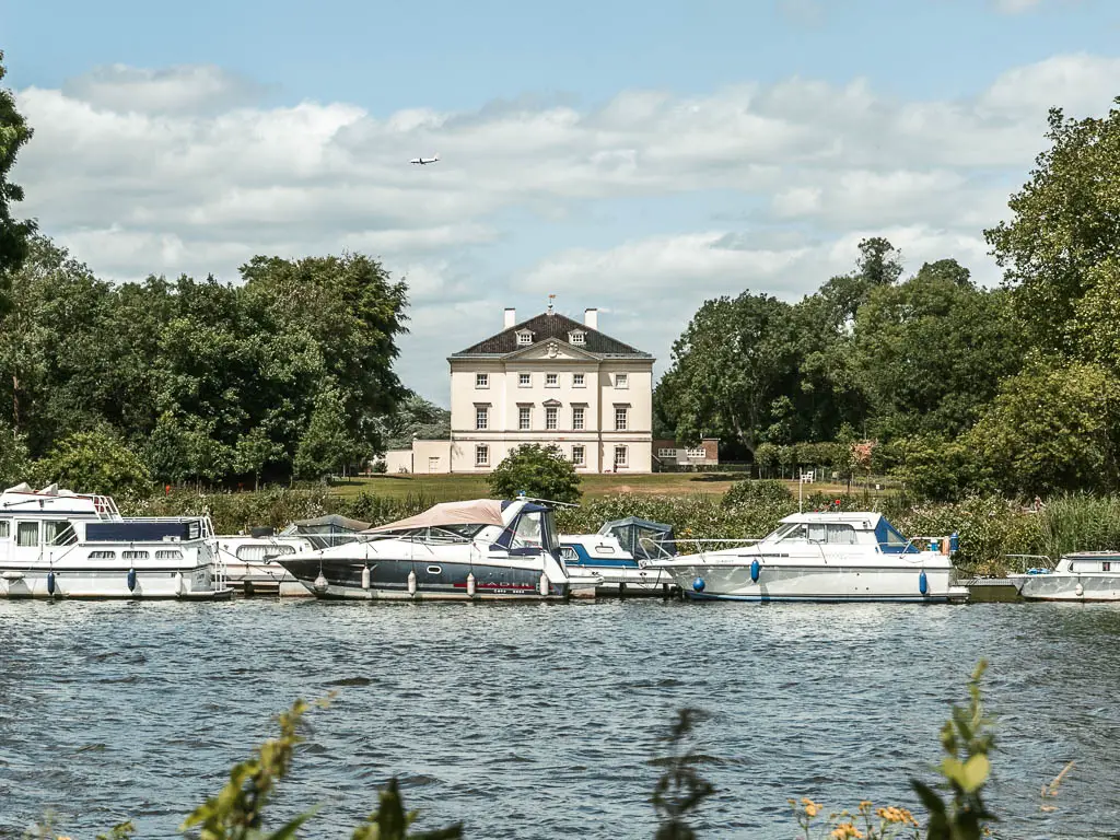 Looking across the river to a green lawn and large box style grand Manor House, partway through the walk between Hampton Court to Putney. There are white boats lining the other side of the river. The house is surrounded by large green leafy trees.