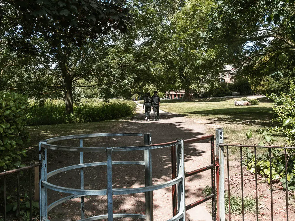 A metal gate leading to a walking oath through the green. There are two people walking along the path ahead.