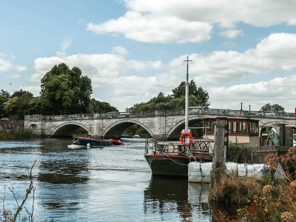 Looking along the river to a grey stone archway bridge along the walk from Hampton Court to Putney. There are a couple of boats in the water.