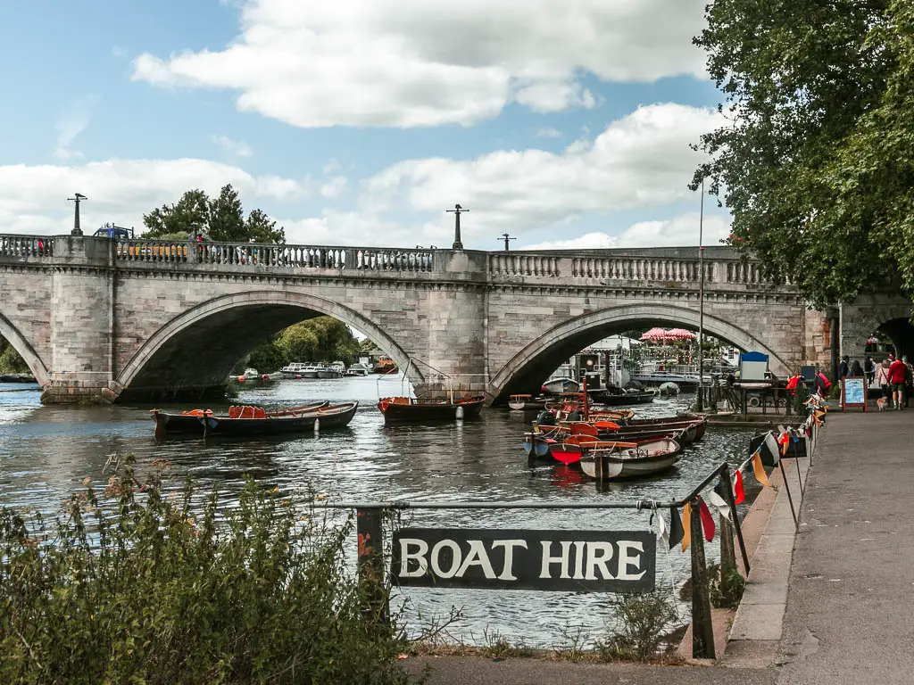 A grey stone archway bridge ahead over the river, with small row boats below it. There is a sign saying 'boat hire'.