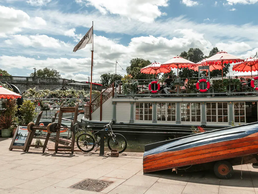 A restaurant boat with sun umbrellas. There is an upside down row boat in front of it on the path, and a railing with a bicycle chained to it.