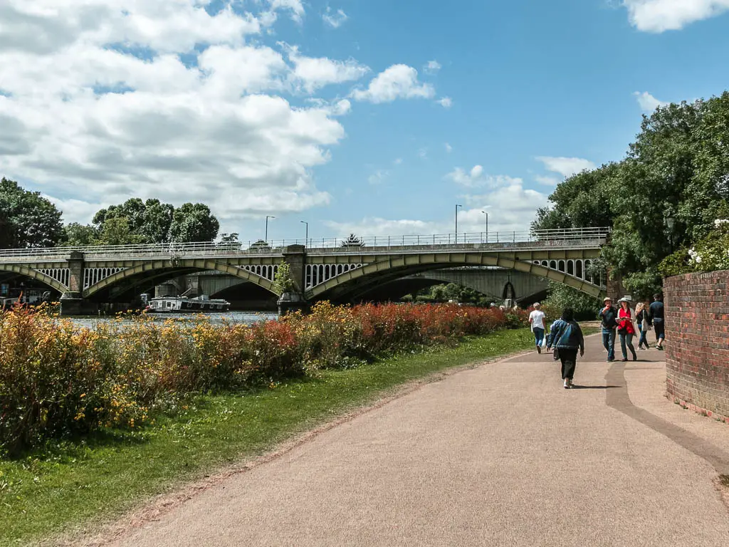 A wide walking path leading to a bridge ahead. The right side of the path is lined with a strip of grass and bushes. There are lots of people walking on the path ahead.