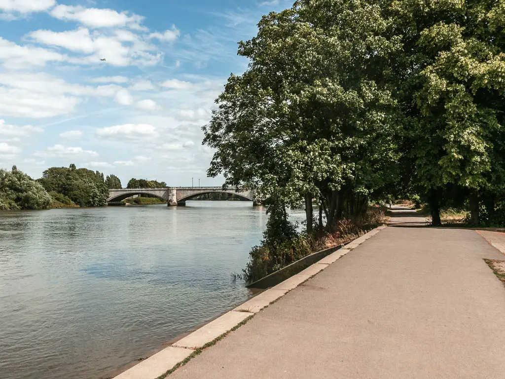 A wide walking path on the right leading towards some trees, with the river to the left and an archway bridge in the distance ahead.