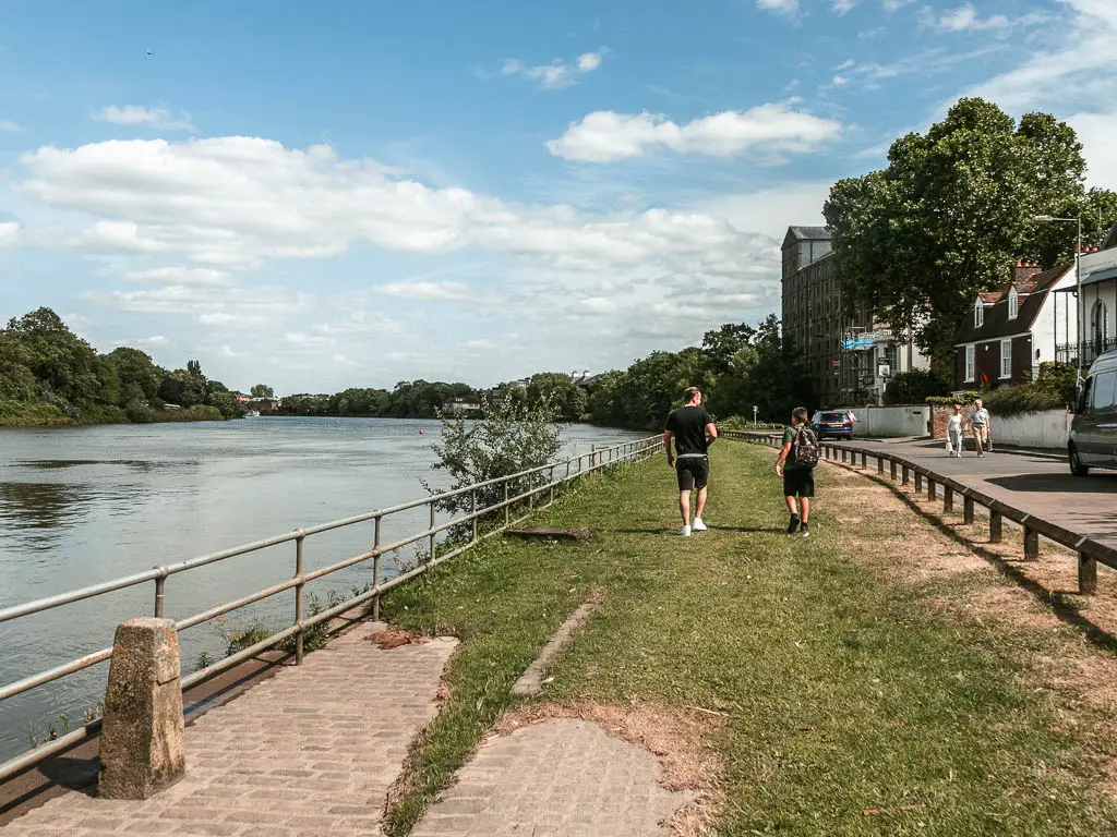 A wide strip of grass leading ahead with the river to the left and road to the right. There is a metal railing between the grass and river. There are two people walking on the grass ahead.