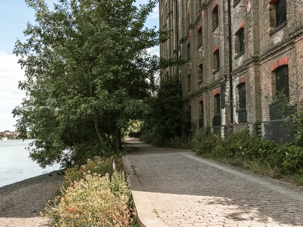 A stone paved path leaden g ahead, with a derelict brick building to the right and river to the left.