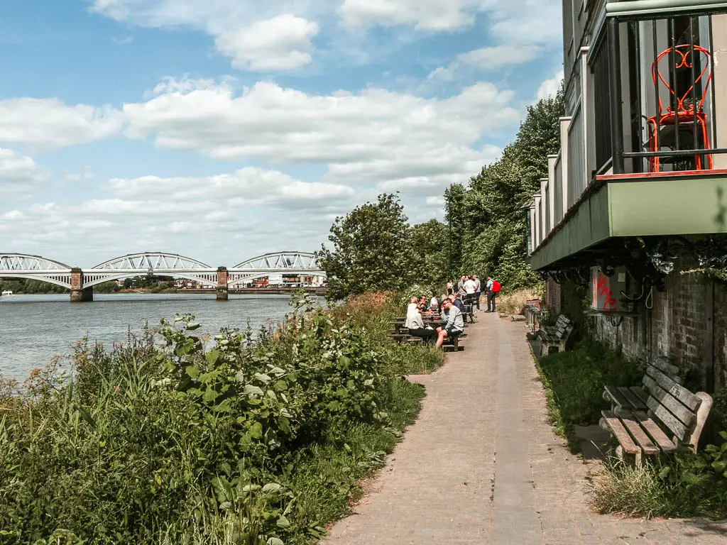 A path leading ahead, with buildings to the right and a strip of tall grass then river to the left. There is a bridge over the river ahead. There are people sitting on picnic benches next to the path ahead.