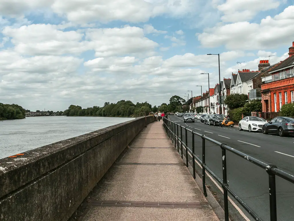 A long straight path with metal railings and road to the right and a stone wall and river to the left. On the other side of the road are houses with cars parked outside.