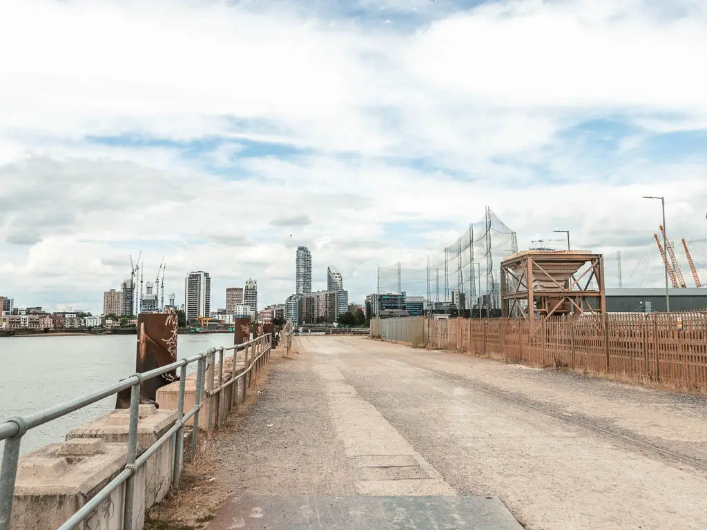 A wide walkway with railings and river to the left, and industrial things to the right.