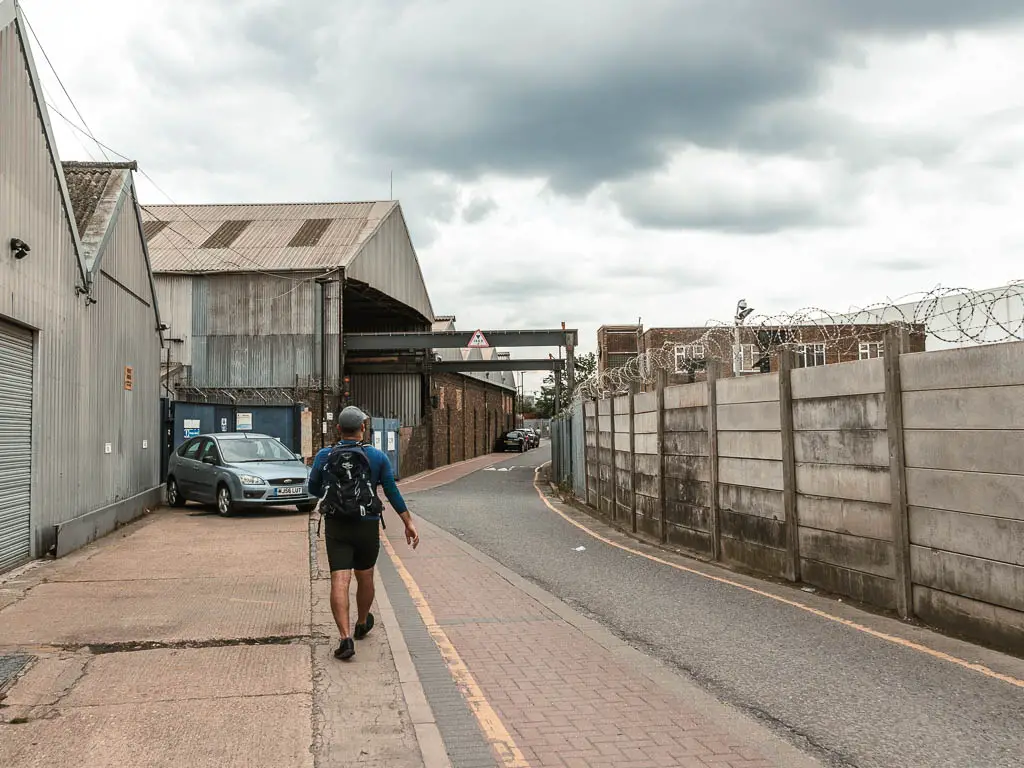 A road with warehouse buildings on the left, a stone wall with barbed wire on the right, and a man walking ahead.