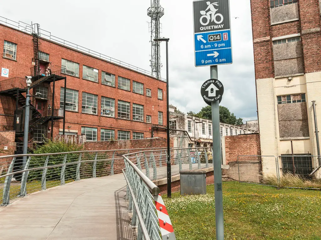 A bridge walkway with factory buildings ahead, and a thames path sign on a pole.