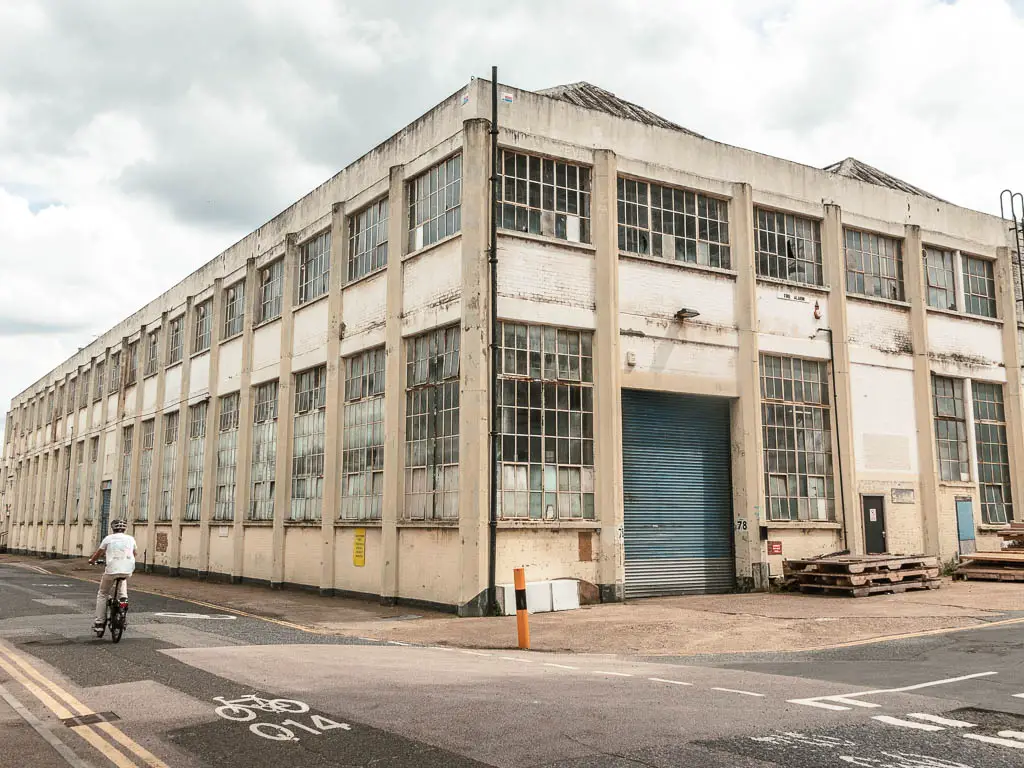 Looking towards the corner of a derelict factory building partway through the walk from Tower Bridge to the Thames Barrier. There are broken glass window panes in the building. There is a cyclist on the road on the left.