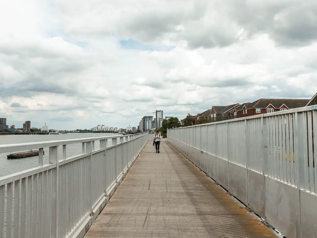 A long straight walking path lined with silver railings. There is a person walking along it ahead, and the river is to the left.