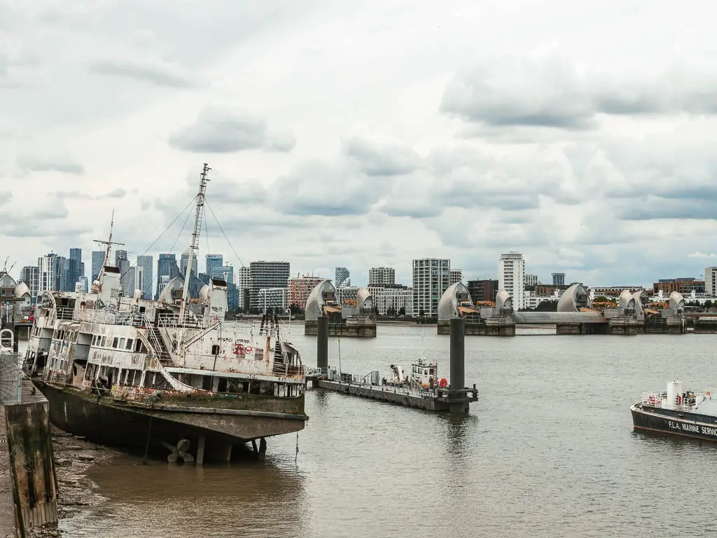 A leaning derelict ship on the left side of the river at low tide, with the Thames Barrier behind, on the walk from Tower Bridge. The skyscrapers of Canary Wharf are visible in the distance.