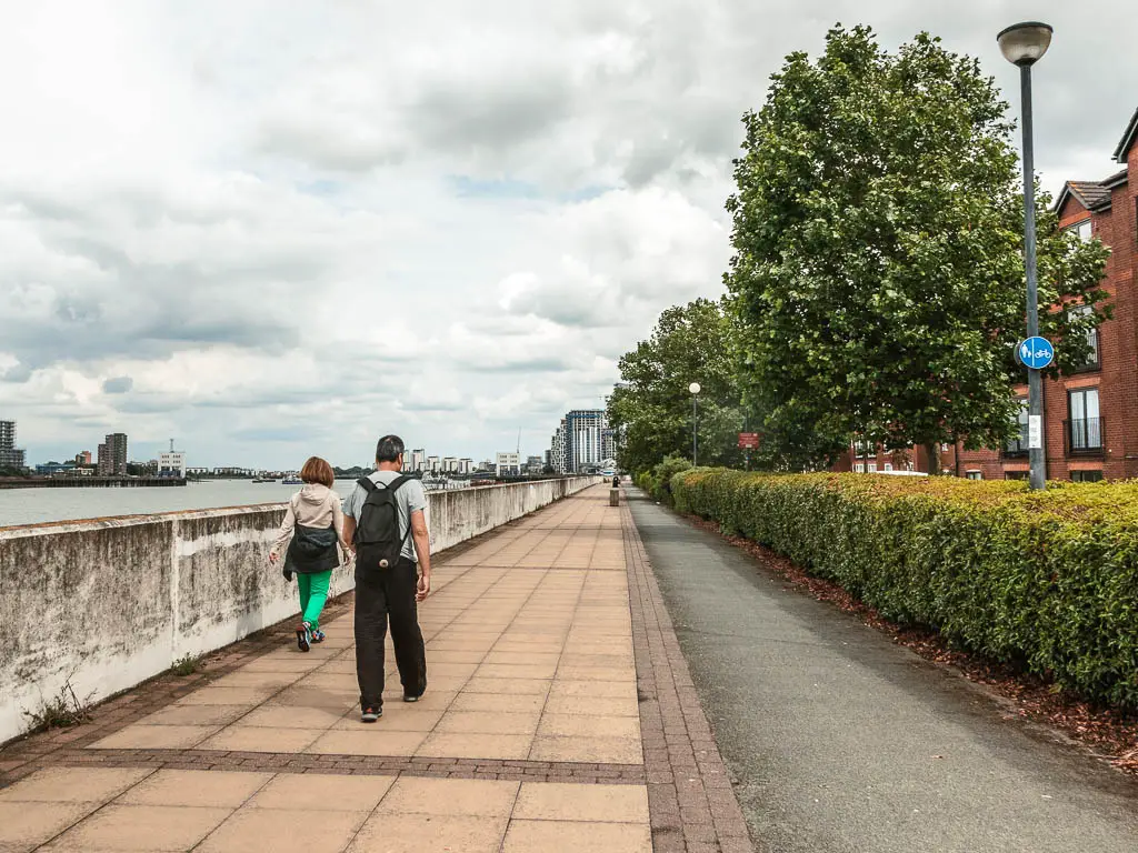 A long straight walking oath with green hedges to the right and concrete wall to the left. There are twp people walking on the path.