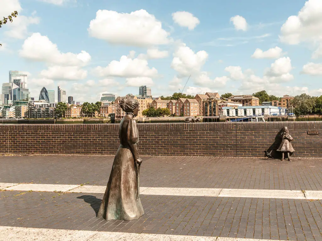 Statues of Dr Salters Daydream on the walkway, lined with a brick wall on the other side. Houses and skyscrapers are visible in the distance.