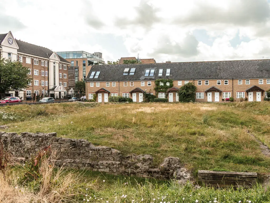 Wall ruins surrounding a grass patch, and a row of terraced houses on the other side.