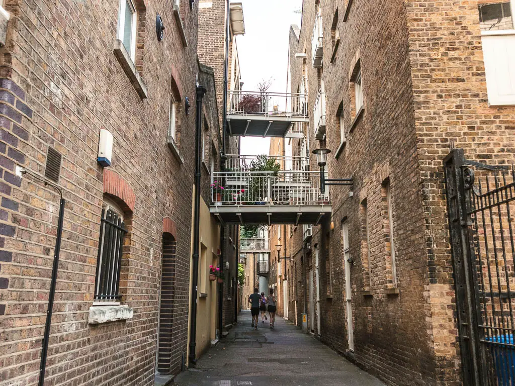 An alley walkway between brick buildings, on the walk between Tower Bridge and the Thames Barrier. There are bridge balconies between the buildings. There are some people walking below.