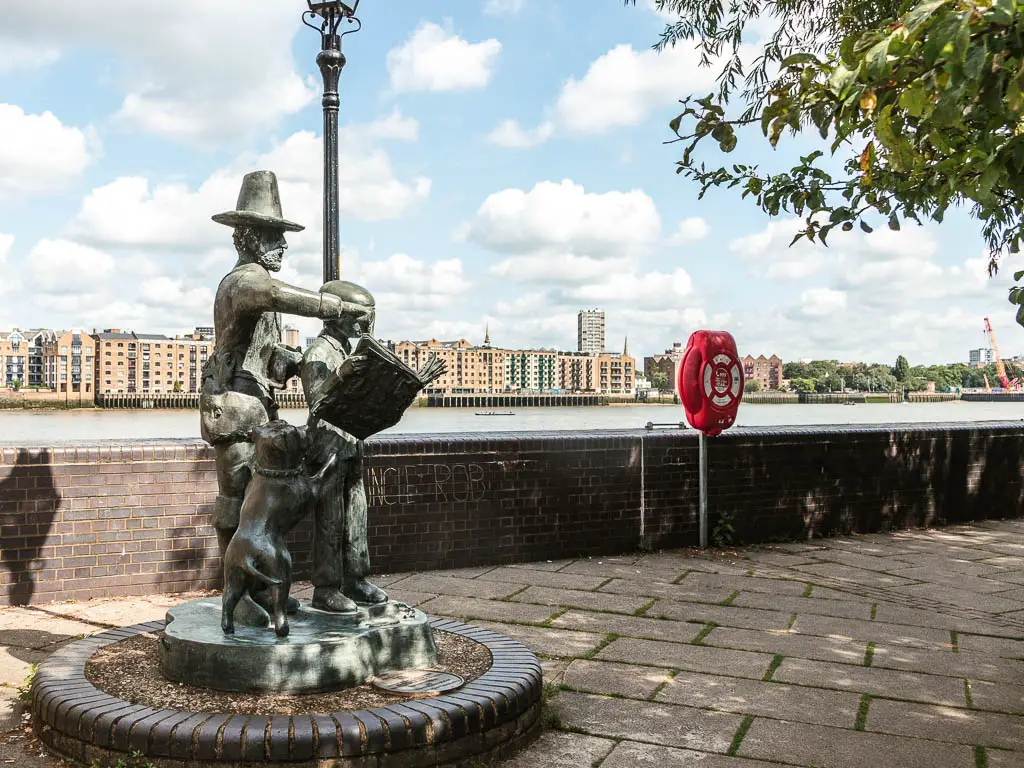 A sculpture of a man, child, and dog o0n the walkway, with the brick wall behind them and river on the other side.