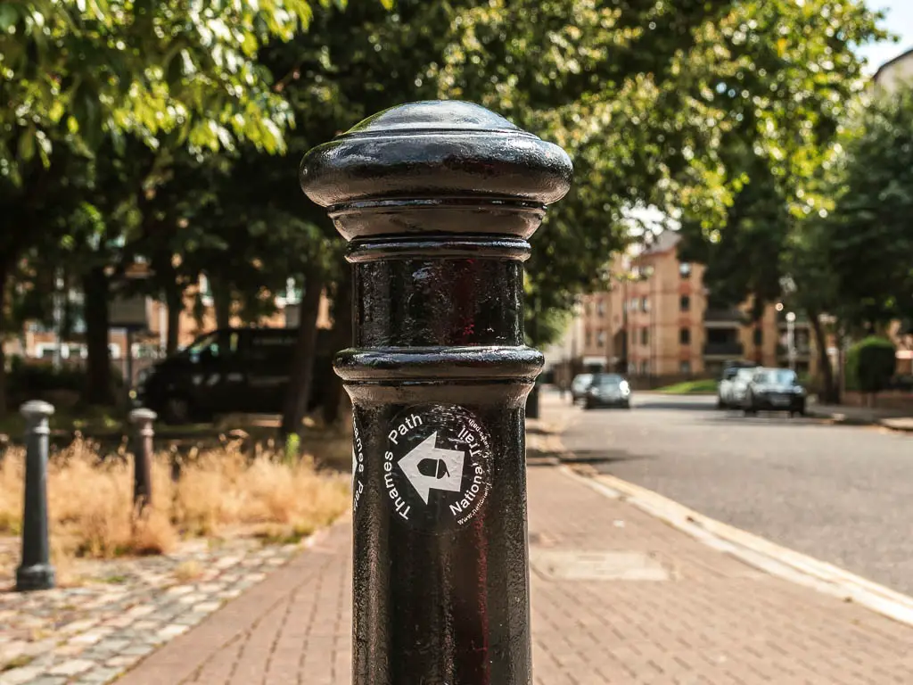 A black thames path sign, on a black post, with white allow pointing left, on the walk between Tower Bridge and the Thames Barrier.