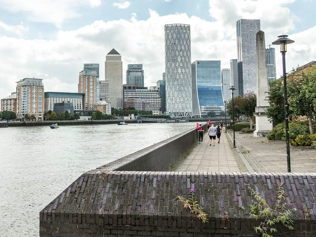 Looking along the walkway with a brick wall to the skyscrapers of Canary Wharf ahead, partway through the walk from Tower Bridge to the Thames Barrier.