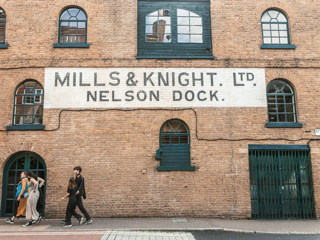 A brick wall with old sign painted in white saying Mills & night. LTD Nelson Dock. There is a family walking next to the wall on the pavement. 