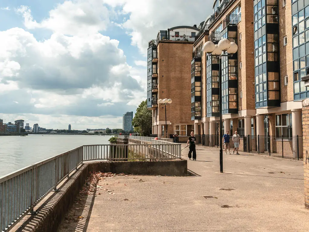 A wide walking path, with apartments on the right, black railing on the left, and river to the left of it. There are a few people walking on the path.