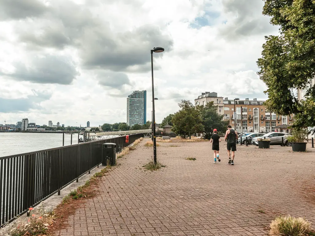 A wide walking path with two people ahead. There is a black railing and river to the left.