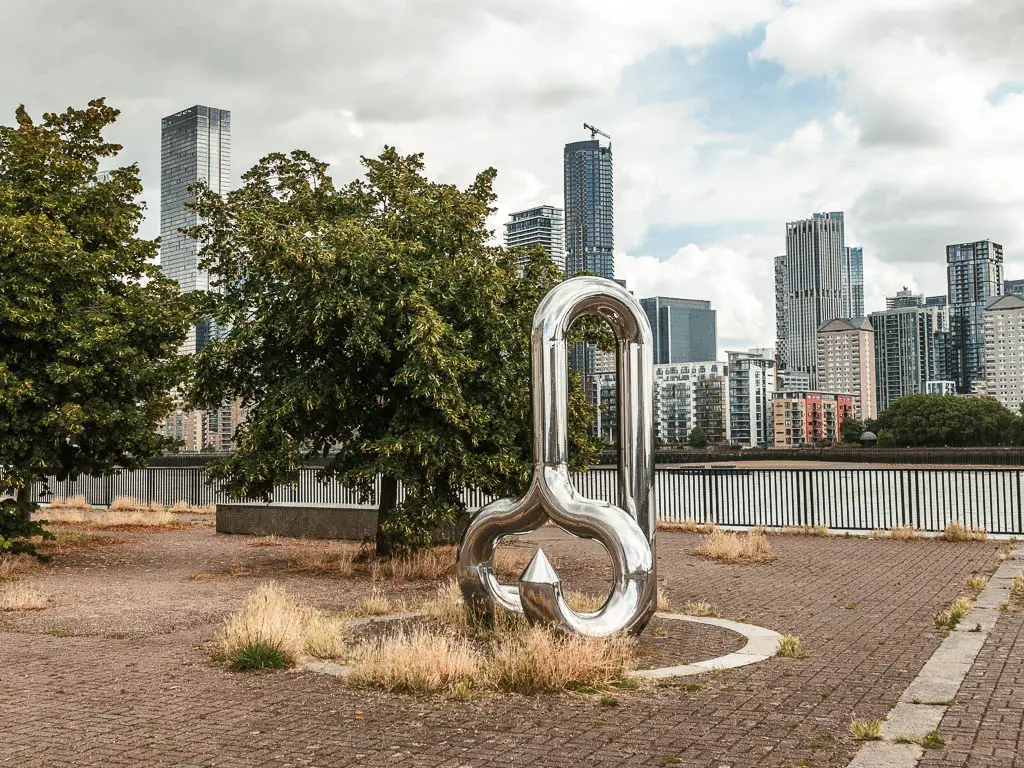 A sculpture on the walkway, when walking from Tower Bridge to the Thames Barrier. There are skyscrapers in the distance, on the other side of the river.