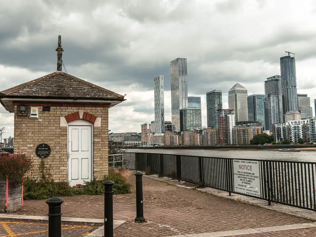 A small brick lock hut on the walkway on the  left, with black railings then river to the right, and the skyscrapers of Canary Wharf on the other side of the river.