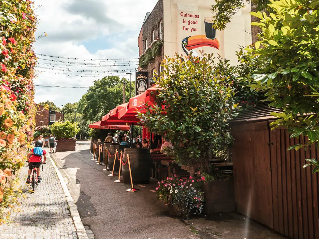 A narrow road with a pub on the right side, with red parasols covering the outdoor eating area.