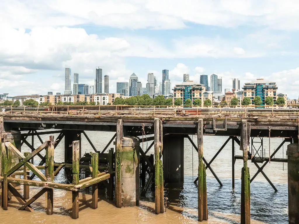 Looking over the pier at low tide with its supports exposed and canary wharf skyscrapers in the distance, on the walk from Tower Bridge to the Thames Barrier.