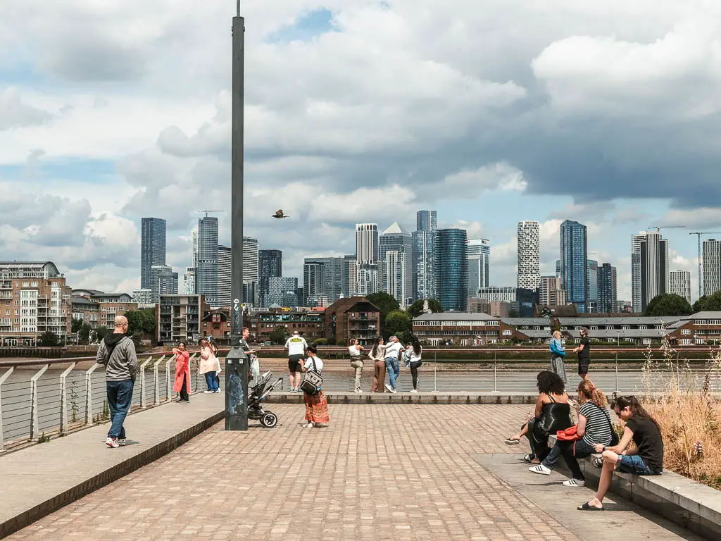 Looking along the walkway to the river and Canary Wharf skyscrapers on the other side, in Greenwich. There are lots of people sitting and walking.