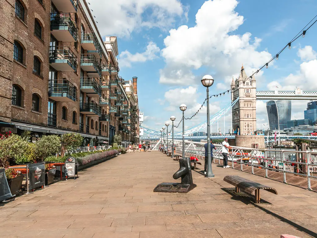 A wide walkway with brown brick buildings to the left, and railings and river to the right. 