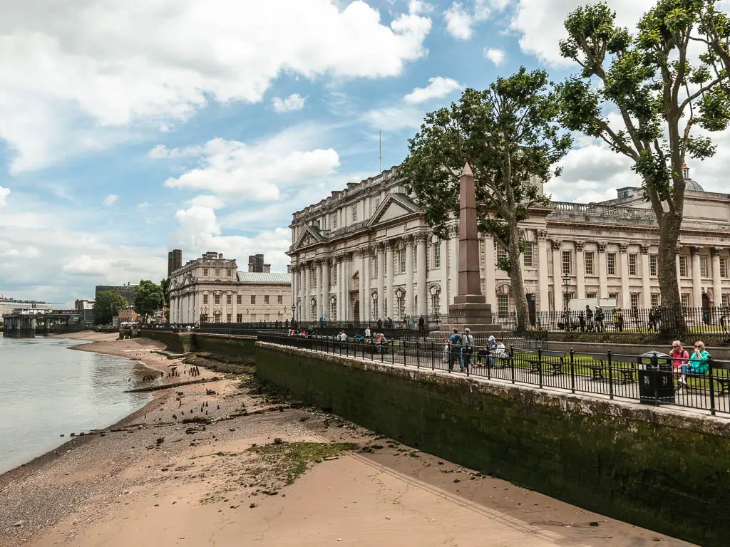 Looking along the sand back of the river at low tide, with a walking path on the other side and grand palatial building, partway through the walk from Tower Bridge to the Thames Barrier.