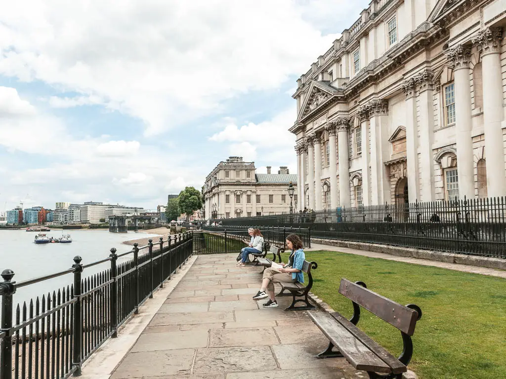 A stone paved walking path with benches and small green to the right, with a grand palatial building on the right side and black railing and river to the left. There are a few people sitting on the benches.