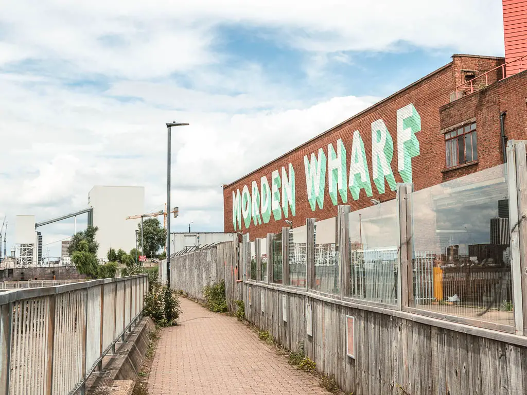 A narrow walkway with railing to the left and brick building to the right behind the wall. The building has 'Modern Wharf' written in bold across it.