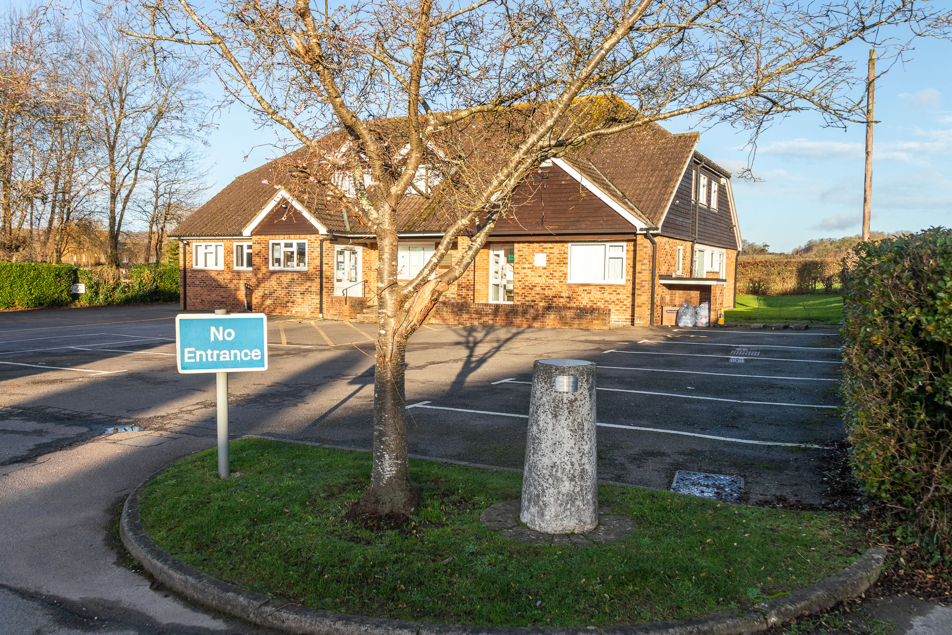 A grey pillar for Saturn sitting on a patch of green next to an empty car park. The GP surgery building is behind. There is also a leafless tree on the green next to the pillar, and a blue no entrance sign.