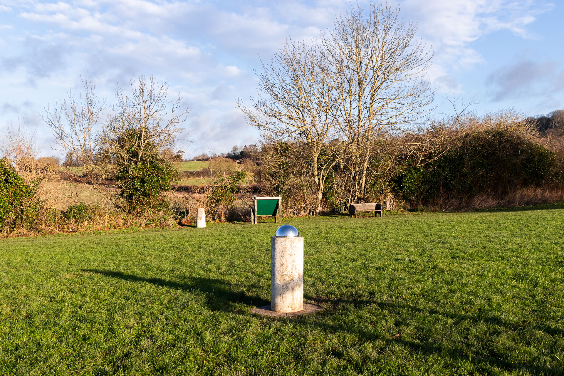 The sun marker for the otford solar system walk. It's a reflective dome on top of a light grey pillar, in the grass field. Past it you can see a green information board with another pillar next to it on the edge of the field. 