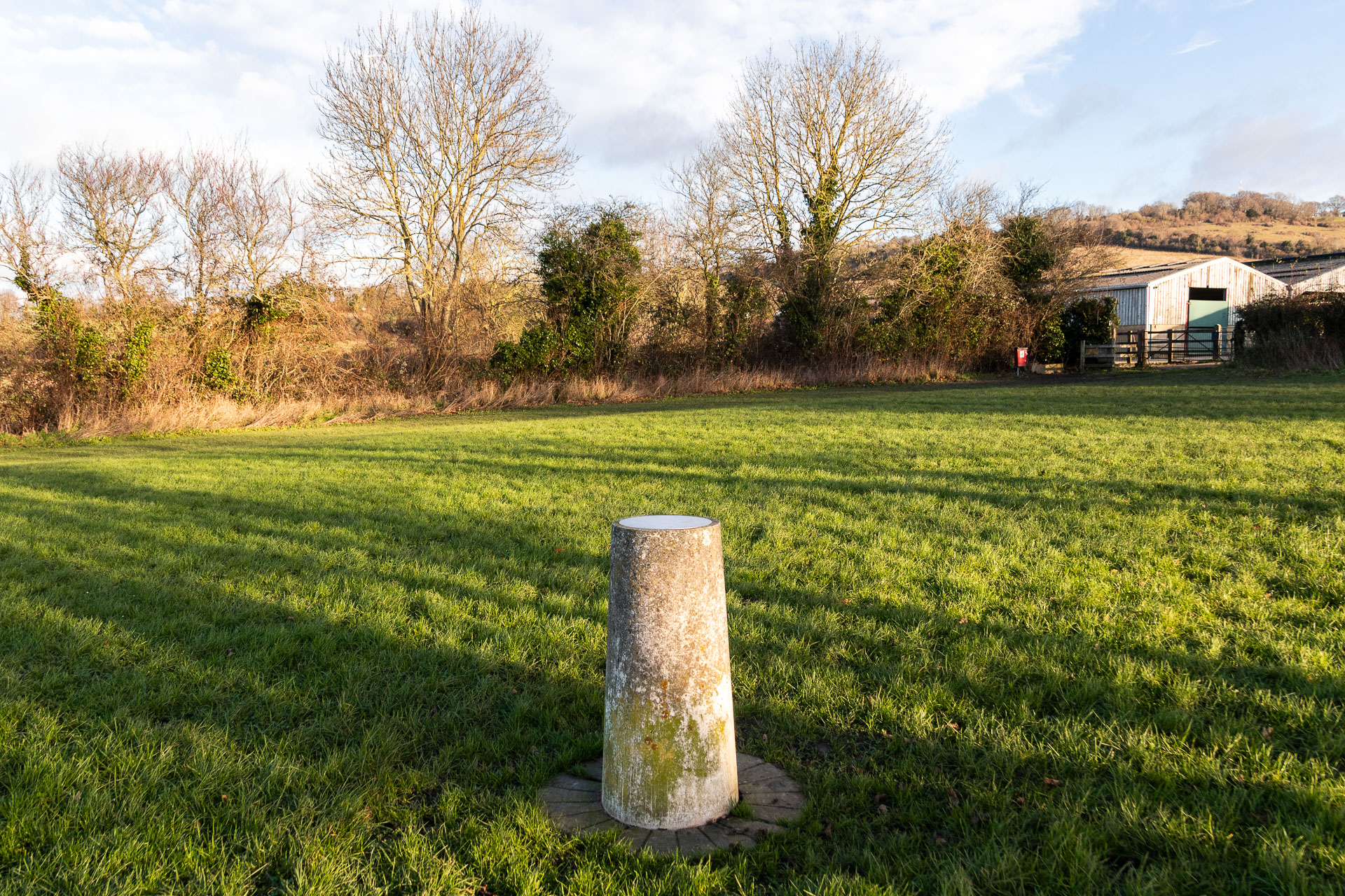 The earth pillar of the Otford Solar System walk, in a green grass field. The sun is shining down with strips of shadow creating lines across the field. The edge of the fill ahead is lined with bushes and trees.