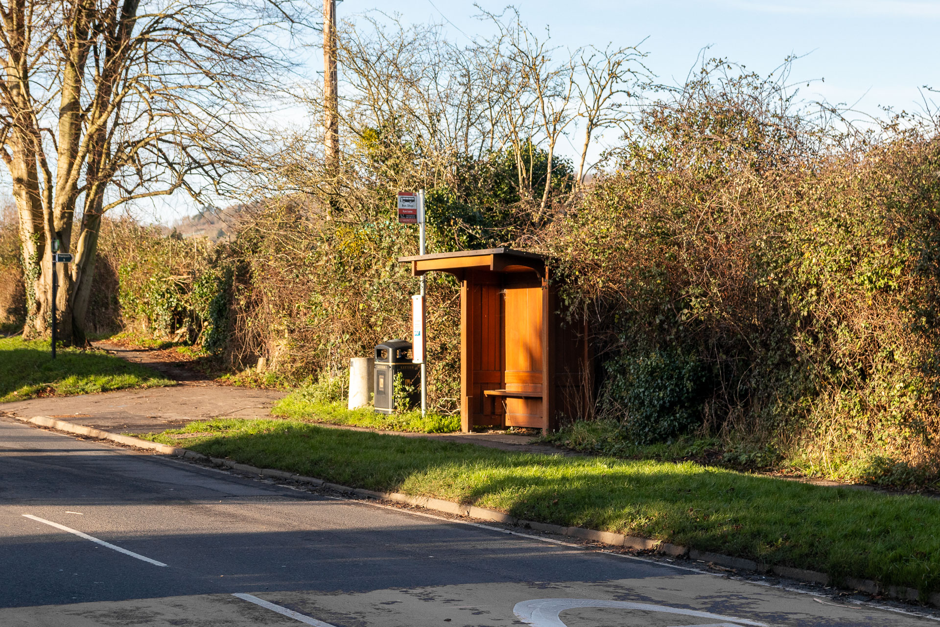 Looking across the road to a wooden  bus stop shelter, with a black bin and grey pillar for Uranus to the left. There is a hedge behind them.