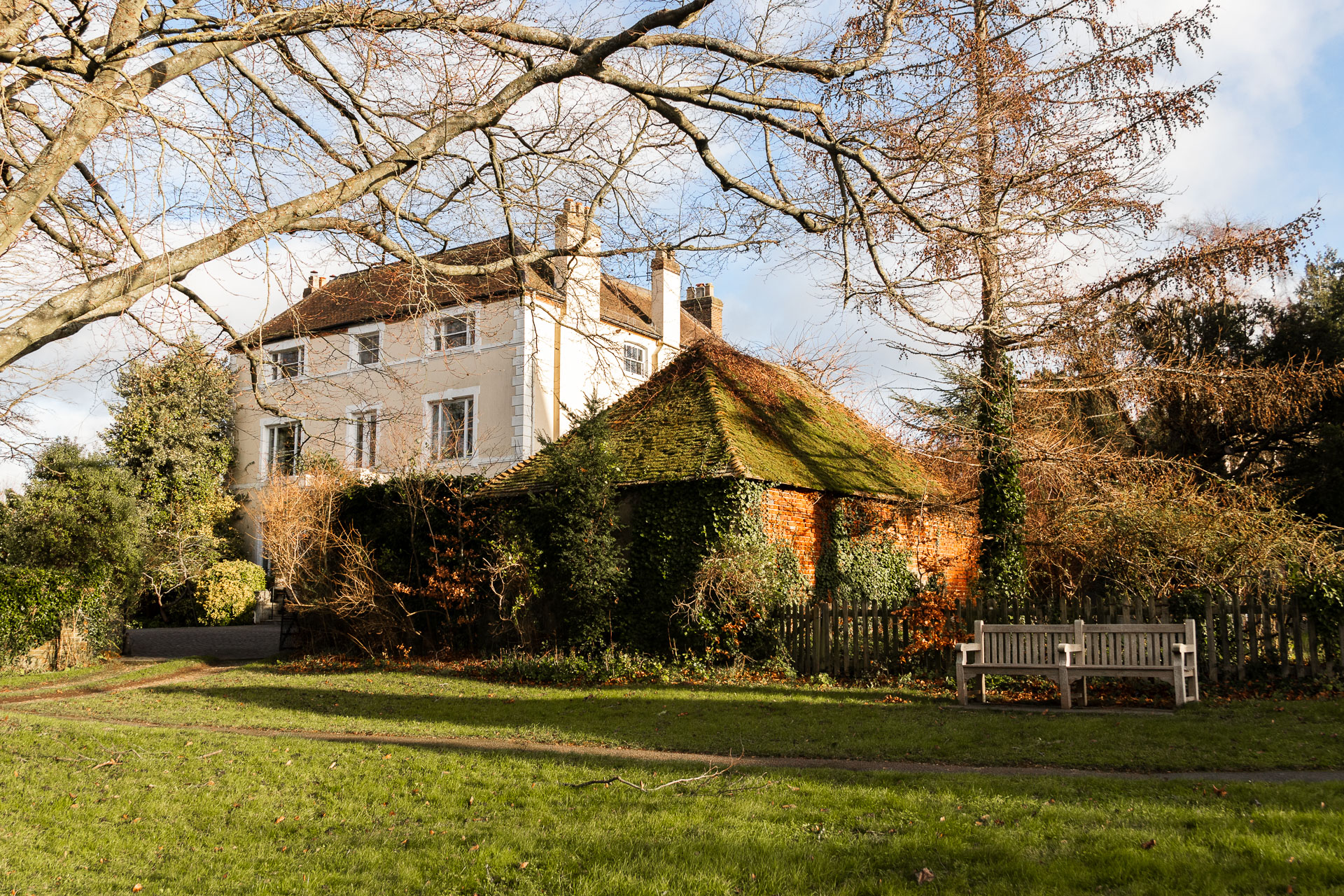 Looking across a neatly cut green to a grand white walled house behind hedges and bushes ahead. There are tree branches coming into frame from the left. There is a wooden bench on the green on the right. 