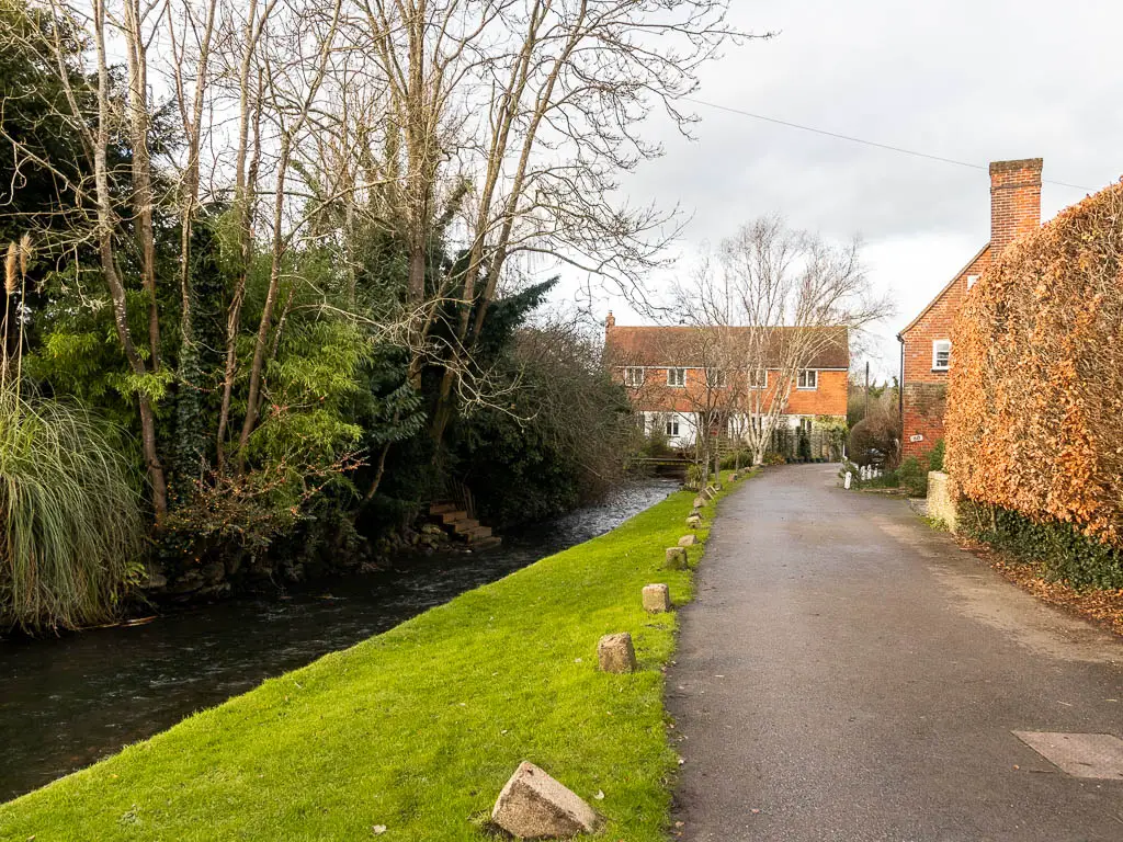 A neat path leaden g ahead, with a strip of green to the left, and a stream to the left of it. The road leads to an orange cottage ahead.