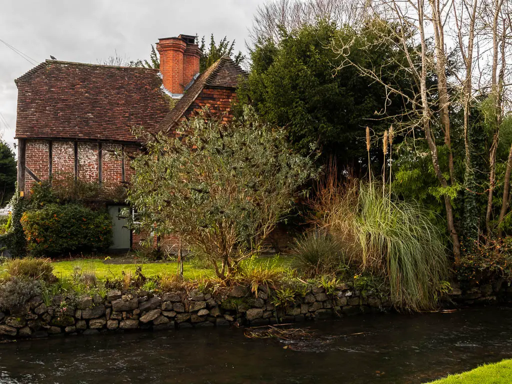 Looking over a steam of water lined with a stone wall, and a green and cottage partially hidden by trees on the other side.