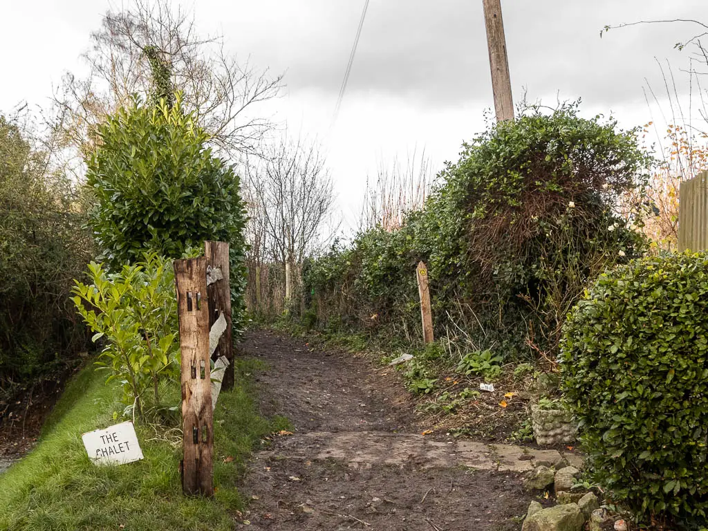 A dirt trail, lined with hedges. There is a wooden fence to the left, and white sign saying 'the chalet'.