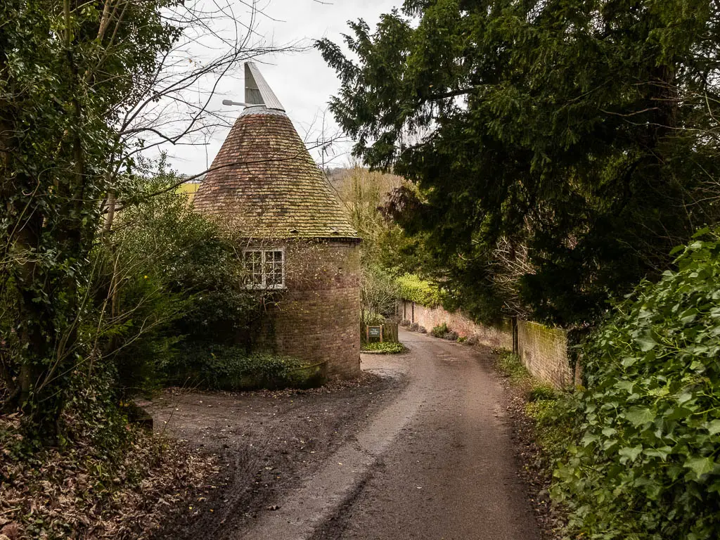 A road leading ahead and giving to the left around an oast house with a cone shaped roof, on the walk from Shoreham to Otford. The road is lined with buses and trees with green leaves.