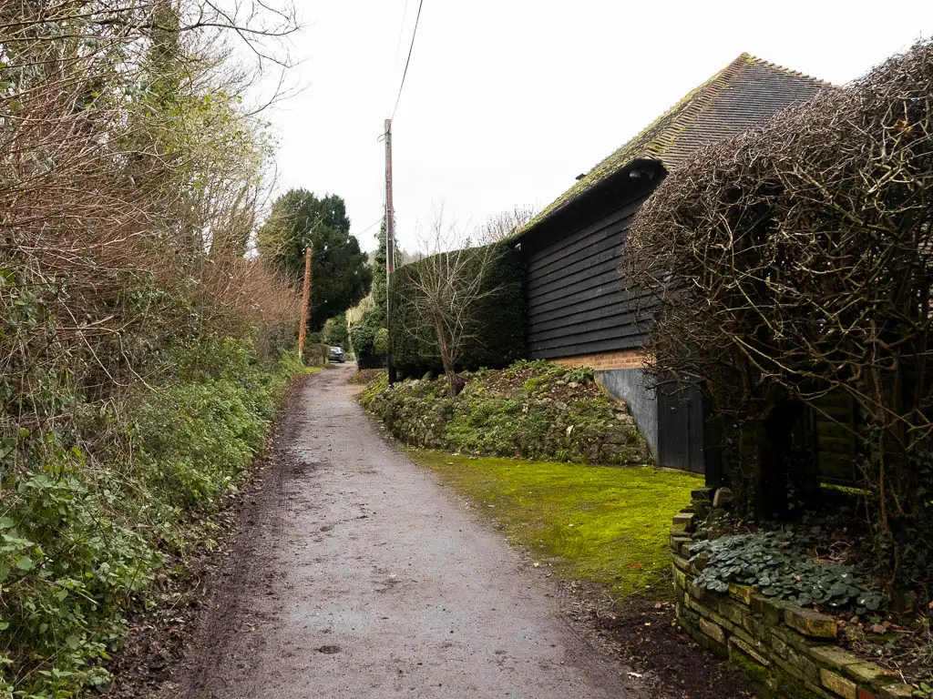 A narrow road leading uphill, lined with hedges to the left and a hedge and black timber framed cottage to the right.