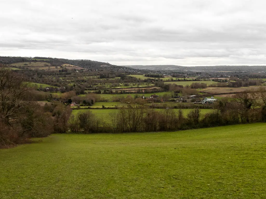 Looking down the green grass hill to the valley below, near the end of the walk from Otford to Shoreham. The valley is filled with a mix of fields, trees, and barely visible cottages. There is a hill on the other side of the valley.